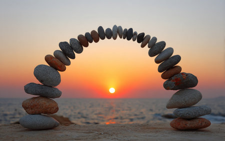 A stone arch is meticulously constructed against a stunning sunset backdrop. The composition presents a balanced arrangement of stones in various colors, shapes, and sizes. Warm hues dominate the scene, with sunlight and soft light enhancing the textures. This image could be used for design, articles, or presentations.の素材