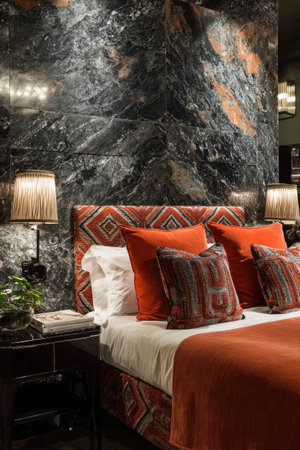 This interior photograph features a bedroom with a large decorative wall behind the bed. The bed has multiple pillows and a red blanket. Table lamps are on either side of the bed. The scene is lit with warm light and shows a high-end interior. Suitable for articles about interior design or hospitality.の素材