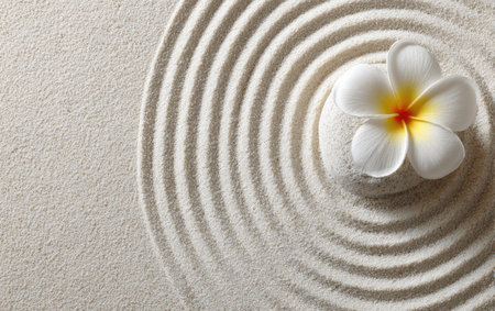 A close-up showcases a frangipani blossom resting on textured, raked sand. The circular patterns contrast with the flower's pristine white petals and vibrant yellow center. The composition, with ample copy space, could be suitable for designs related to wellness, spa treatments, or meditative practices.の素材