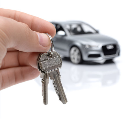 A hand presents keys with a silver vehicle blurred in the background against a white backdrop. The image uses a shallow depth of field to emphasize the keys. The scene uses neutral tones, suitable for various commercial uses. The composition highlights concepts such as ownership and transportation.の素材