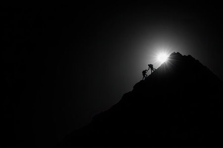 Two figures climb a rocky mountain against a bright, radiant sun. The high-contrast image displays a dark silhouette against a bright background, emphasizing determination. The composition creates a dramatic effect. This picture is suitable for concepts of teamwork and challenge.の素材