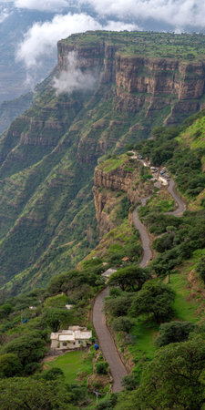 An elevated perspective reveals a steep mountain with a road meandering across the terrain. The scene showcases an abundance of green vegetation covering the slopes and cliffs. Cloudy skies hover above the summit. This visual may be suitable for environmental, travel or tourism-related projects.の素材