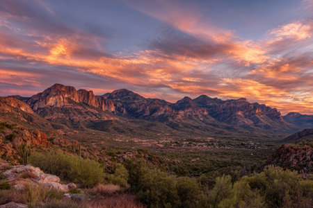 A stunning mountain range dominates the frame under a dramatic sky filled with fiery orange and purple clouds. The landscape features varied terrain and textures, with distant peaks and lush green vegetation in the foreground. This vista could be ideal for various visual projects related to nature, travel, or environmental topics.の素材
