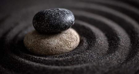 Two smooth stones are balanced on a bed of textured sand, creating a zen-like composition. The image features a contrast of light and dark colors, with attention drawn to the round shapes of the stones and the surrounding patterns. This visual can be used for various purposes, including wellness and meditation.の素材