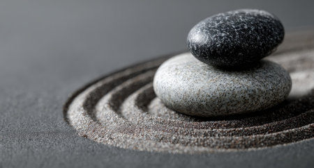 Two stones are stacked on a bed of textured sand, creating a balanced composition. The image showcases contrasting textures with soft gray and black hues. The lighting appears to be diffused, suggesting an indoor setting, suitable for use in wellness or mindfulness content.の素材