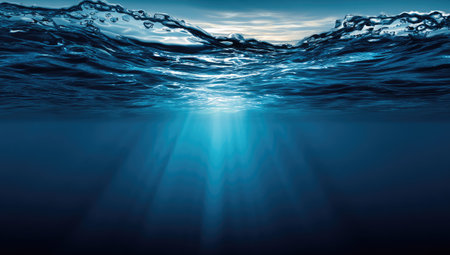 This image depicts an underwater perspective with the water surface visible above. Sunlight penetrates the water, creating beams of light. The composition features shades of blue. This image could be used for projects related to nature, science, and design.の素材