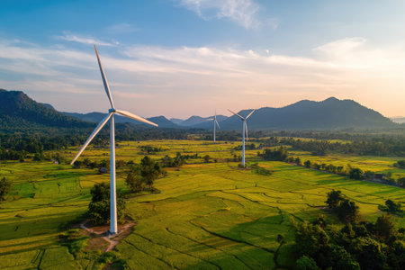 This image showcases several wind turbines standing tall in a field with a backdrop of rolling hills. The scene is lit by the soft glow of either sunrise or sunset, with the turbines casting long shadows. The style is a professional and captures the essence of green energy and environmental consciousness. Suitable for various commercial purposes, including articles, and presentations.の素材