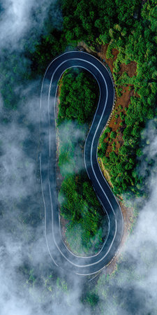 An aerial perspective showcases a winding road slicing through a vibrant green mountain landscape. The road curves gracefully, suggesting a journey through nature. The composition features rich green vegetation, hints of brown earth, and wispy clouds. It offers visual potential for various applications, including travel, environment, and transportation themes.の素材