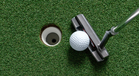 A golf ball is positioned near a putting hole. A golf club rests nearby. The scene displays a close up view on a green surface. The artificial grass provides a texture for the sport environment, illuminated with natural daylight. Suitable for commercial and editorial uses.の素材