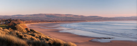 A scenic coastal image showcases a sandy beach meeting the ocean under a soft, pastel-colored sky. The composition includes sand dunes in the foreground, leading to a shoreline with gentle waves. This landscape photography could be used for environmental themes or to convey natural beauty for various commercial applications.の素材