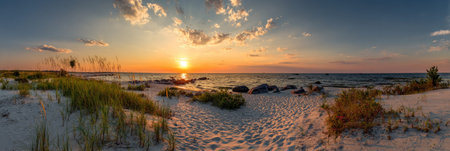 A panoramic view displays a vibrant sunset over a sandy beach, with the sun reflecting on the ocean water. The image showcases a dramatic sky with clouds and various shades of orange and blue. This photograph is suitable for different commercial purposes, including advertising or editorial content.の素材