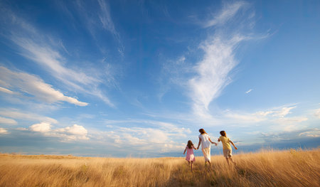 A family is depicted running through a vast field of golden grass, holding hands. The image showcases a blue sky dotted with white clouds, creating a bright and airy atmosphere. The composition features a low angle, soft lighting, and a sense of movement, suitable for various commercial and lifestyle projects.の素材