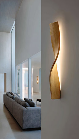A wooden wall lamp is the focal point, showcasing a curved design. The lamp emits warm light against a white wall. A modern interior reveals a sofa and an open space, suggesting architectural photography. The image could be used for interior design, home decor, or lifestyle projects.の素材