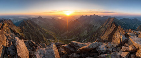 A wide shot presents a mountain range at dawn, bathed in golden light. Jagged peaks and rocky foreground elements frame a distant view. The composition employs natural color and texture, depicting a vast open environment. It could be used to illustrate concepts related to travel or scenic beauty.の素材