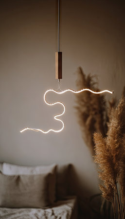 An artistic light fixture is suspended from the ceiling, emitting a soft, warm glow. Its minimalist design features a wooden element and a wavy light shape, standing out against a blurred background. The scene includes dried plants and a beige color palette, implying an indoor setting. This image could be used for interior design or home decor.の素材
