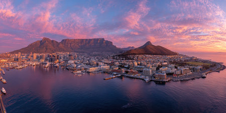 A panoramic view captures a coastal city nestled between mountains and a tranquil ocean. The sky displays vibrant hues, including pinks, oranges, and blues. Buildings and natural elements merge to create a stunning visual. The image may be used for travel, tourism, or environmental purposes.の素材