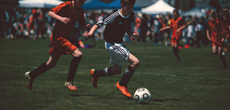 Players are actively engaged in a soccer game on a green field. The image exhibits a dynamic composition with a focus on action, featuring a ball and players in motion. The attire consists of sports uniforms, set against the backdrop of a sunny day with other participants. Suitable for sports, recreational or team related visual content.の素材
