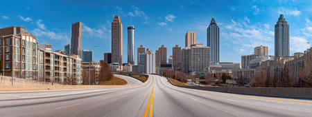 A road stretches towards a cityscape of modern high-rise buildings under a clear, blue sky. The composition utilizes a perspective with leading lines to emphasize depth. The style presents a realistic depiction with natural lighting. Suitable for various commercial applications such as websites, advertising, and print media.の素材