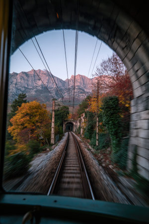 View from a moving train reveals tracks leading into a tunnel, framed by a window. The scene includes mountain range, autumn trees, and overhead wires. The composition features natural lighting. Suitable for illustrating travel, transportation, and scenic landscapes.の素材
