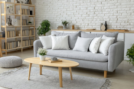 This image showcases a modern living room featuring a gray sofa adorned with white and gray pillows. A wooden coffee table sits in front of the sofa, and plants add a touch of nature. The room is illuminated by natural light, with a neutral color palette and a white brick wall. Suitable for home decor or lifestyle content.の素材