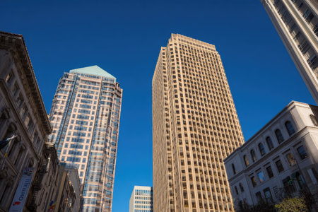 The image showcases multiple skyscrapers and buildings under a clear, vibrant blue sky. The architectural styles vary, with some structures featuring traditional facades and others displaying modern glass exteriors. The composition uses a low-angle perspective, emphasizing the height of the buildings and the open space. The scene is illuminated by natural light and could be suitable for various commercial applications.の素材