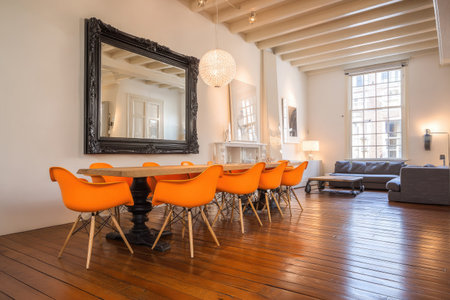 A spacious dining area features a wooden table and bright orange chairs, illuminated by warm lighting. A large, ornate mirror dominates the wall, reflecting the room's elegant design. The space showcases a clean, modern style with a focus on open space, suitable for various commercial applications.の素材