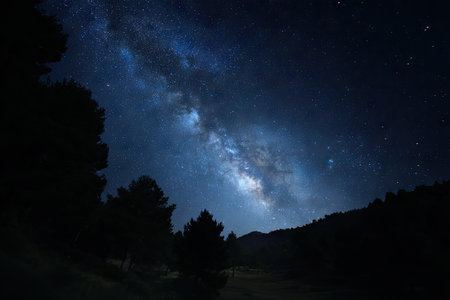 A dark, night sky dominates this image, showcasing the Milky Way galaxy in a stunning display of celestial bodies. Silhouetted trees and a distant mountain range define the earthly landscape. The deep blues and blacks create a dramatic visual, lending itself to editorial and commercial applications.の素材