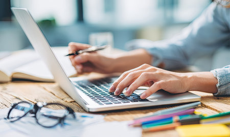 Close up shows hands using a laptop with other items like a pen, pencils, and glasses on a wooden desk. The scene is well-lit, with a shallow depth of field, and the person's arms and hands are in focus. It could be suitable for business or technology-related illustrations.の素材