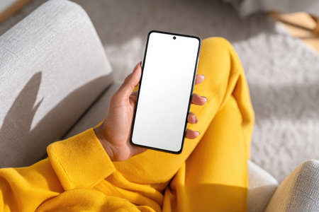A person's hand holds a smartphone with a blank display. The image showcases a close-up view, featuring the device against a backdrop of a yellow garment and a neutral-toned couch. Soft natural light suggests an indoor setting, and the composition provides ample copy space for potential advertising or digital content.の素材