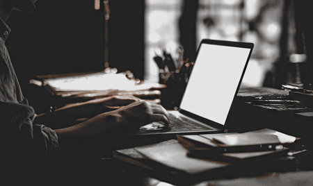A person uses a laptop computer in a low-lit room. The image focuses on the hands and the illuminated screen. Various stationary objects are subtly placed around the desk. The composition employs deep shadows and highlights, suitable for creative projects and website design.の素材