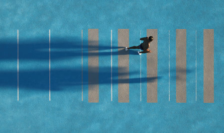 An overhead view showcases a person walking across a crosswalk. The scene is brightly lit, suggesting daytime conditions. The composition features a linear design with shadows and lines creating depth and interest. Suitable for use in projects related to transportation, safety, or urban design.の素材