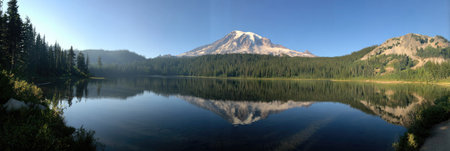 A panoramic view captures a mountain reflected in still water, under a clear blue sky. Lush green trees line the shores of the water, contributing to the overall serene environment. This landscape could be used for editorial and commercial projects depicting natural beauty and the outdoors.の素材