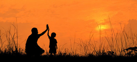 A silhouette depicts a parent and child interacting during a vibrant sunset. The warm orange and yellow hues of the sky contrast with the dark figures. Tall grasses are visible in the foreground. The composition suggests outdoors, possibly during evening. This imagery could be used for editorial or commercial projects.の素材