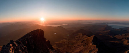 The image captures a mountain ridge at sunrise, bathed in warm golden light. The composition features a panoramic view with a vast sky and layered mountain ranges. The photograph showcases natural textures and a high-angle perspective. Suitable for various commercial applications and illustrating concepts of nature.の素材
