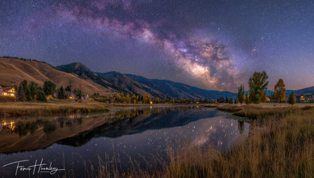 A captivating night landscape showcases a breathtaking view. The image features a body of water reflecting the vast Milky Way galaxy. Rolling hills and mountains frame the scene. The composition utilizes natural colors and lighting, lending itself to possible commercial and editorial applications.の素材