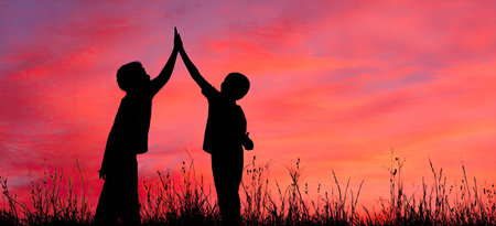 Two figures are silhouetted against a vibrant, colorful sky, engaged in a high-five gesture. The composition emphasizes a bright sunset, with hints of grass. The overall image conveys the feeling of community and achievement, suitable for various illustrative purposes.の素材
