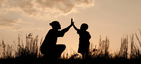 This silhouette image depicts a parent and child high-fiving each other. The composition uses a low angle, showcasing the figures against a warm, orange sky. The figures are dark and solid. The image is suggestive of family interaction, suitable for editorial and marketing materials.の素材