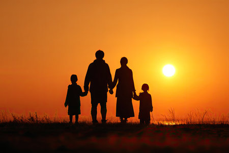 A family of four is silhouetted against a vibrant orange sunset, holding hands. The image features strong backlighting, emphasizing the figures' forms. The composition evokes feelings of warmth and togetherness. Suitable for illustrating themes of family, relationships, and emotional connection; ideal for various editorial and commercial applications.の素材