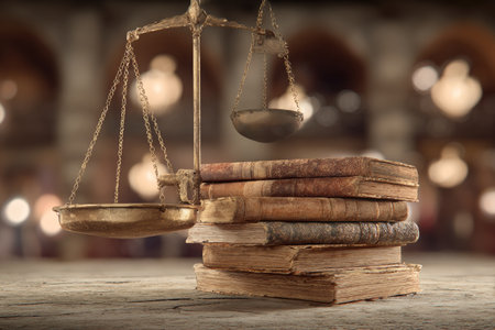 A vintage balance scale stands near a stack of old books, suggesting themes of law, justice, and legal proceedings. The composition features warm tones and soft lighting, hinting at an indoor setting. The image could be used for various commercial and editorial purposes related to law, education, and historical themes.の素材