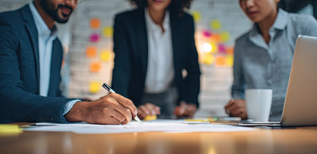 Three business people are gathered around a table, engaged in a collaborative project. The scene showcases hands writing on paper, with a laptop and a coffee cup on the table. The lighting is soft, and the composition suggests an indoor setting. This image could be useful for presentations or commercial purposes.の素材