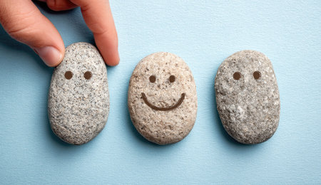 A hand gently places a small rock with a smiling face among two others. The rocks are gray and speckled, set against a soft light blue backdrop. The composition is simple, well-lit, and clean. Potential uses include articles related to mental health, positivity, or team building.の素材