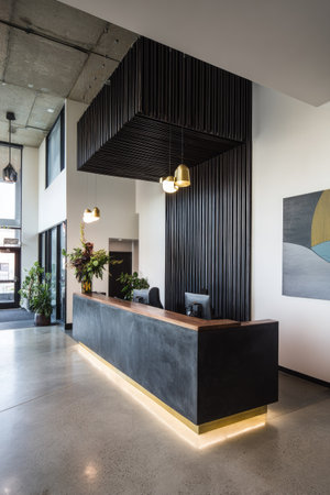 A contemporary reception area features a concrete desk with warm lighting. Dark vertical paneling contrasts with a light colored wall. The space is illuminated by pendant lights, and includes floral arrangements. Suitable for business, office, or interior design applications.の素材