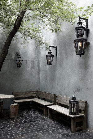 The image shows an outdoor patio setting featuring classic lanterns mounted on a textured wall. A wooden bench and table are present, suggesting a place for relaxation. The composition uses natural light, highlighting the details and creating a peaceful ambiance. It can be suitable for lifestyle, design, and architectural content.の素材