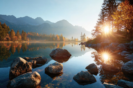 A tranquil landscape depicts a lake surrounded by rocky shores and vibrant autumn foliage. The scene is bathed in warm sunlight, creating reflections on the water's surface. The composition showcases a mountain range in the background, suggesting a natural environment suitable for editorial and commercial applications.の素材