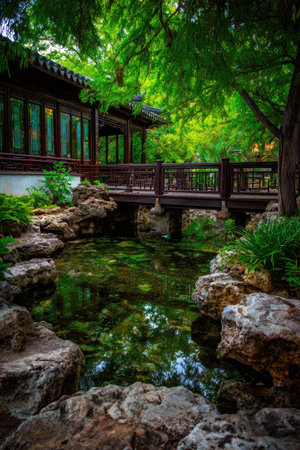 An idyllic scene showcases a traditional Asian garden featuring a wooden bridge spanning a tranquil pond. The image displays vibrant green foliage and a quaint building. The composition is well-lit, emphasizing the natural elements. Suitable for various editorial and commercial uses, this image captures the essence of peace and tranquility.の素材