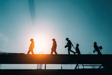 This image shows construction workers silhouetted against a bright, natural light source. The workers are walking on a beam, their forms outlined by the backlighting. The scene features high contrast, with deep shadows and a vivid sky. Ideal for illustrating concepts like teamwork and construction, it can be used for various commercial projects.の素材