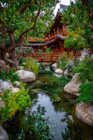 An image showcases a traditional wooden structure with a bridge over a calm water body. Lush greenery surrounds the architecture with various shades of green and brown. The composition uses natural lighting to highlight the textures of the building, rocks, and foliage. This serene view is ideal for travel and landscape-related projects.の素材