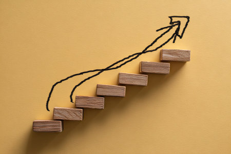 Arrangement of wooden blocks forming steps with an upward arrow drawn above. The composition utilizes a warm, yellow background and natural wood tones. The image uses overhead lighting and offers a simple, graphic style. Suitable for illustrating business concepts, growth, or progress in various commercial contexts.の素材