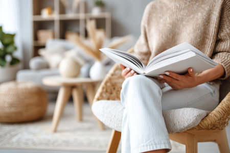 A person is seated in a woven chair, engrossed in reading a book with open pages. The setting appears to be an indoor space with soft lighting and neutral tones. The composition shows details like a sweater, pants, and chair textures suggesting a comfortable environment for activities such as reading. Suitable for editorial and commercial use.の素材