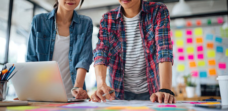 Two people are working together at a desk with a laptop and various papers. The scene is brightly lit, with a modern, casual aesthetic, utilizing primary colors. A wall features numerous colorful sticky notes suggesting brainstorming and planning. This image could be used in business, marketing, or education contexts.の素材